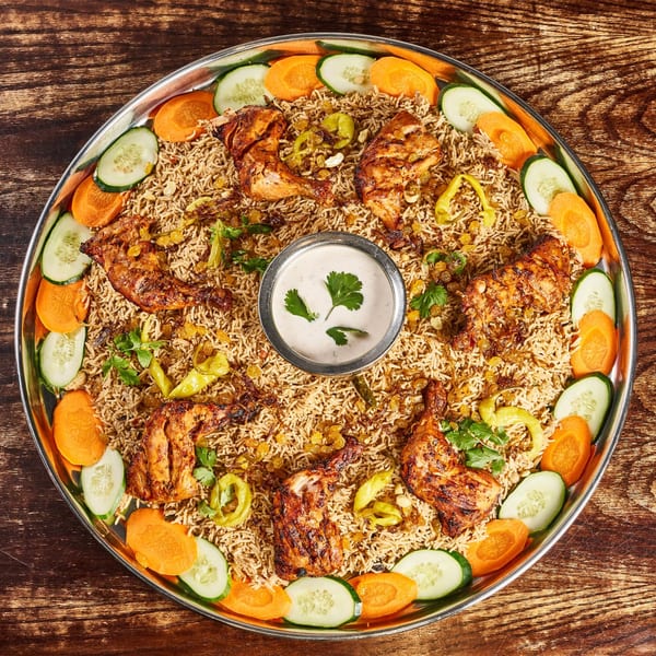 A large platter of Indian chicken biryani with rice, chicken pieces, carrots, cucumbers, and a raita dip.