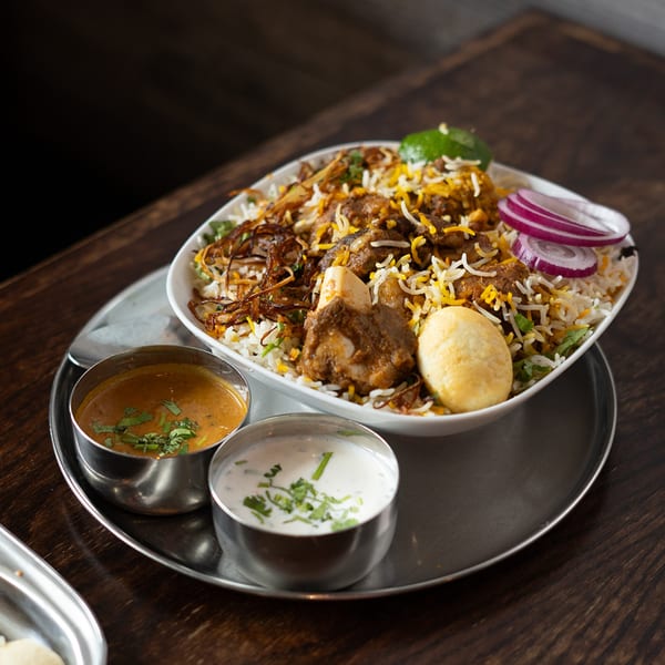 A large bowl of Indian biryani with meat, rice, egg, and red onions, served with two sauces.