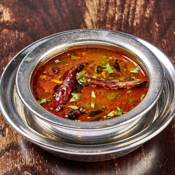 A close-up of a silver bowl filled with vibrant orange-red Indian curry, garnished with green herbs and dried chilies.