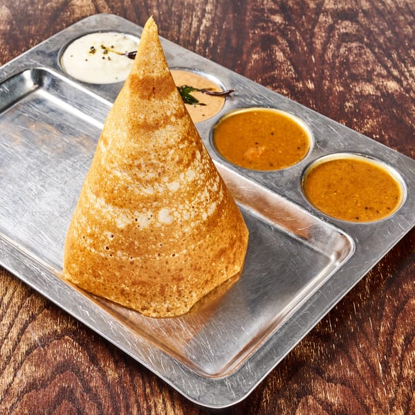 A golden-brown cone dosa with three small bowls of chutney and sambar on a metal tray.