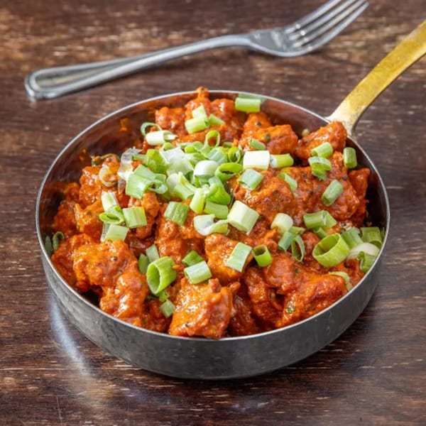 Indian chicken dish in a metal pan, garnished with chopped green onions, on a wooden table.